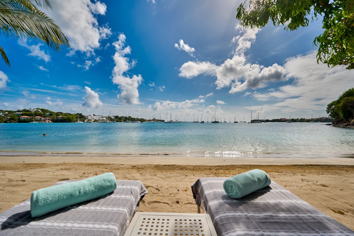 Two loungers with rolled towels sit on the sand at Calabash Hotel's beach, facing turquoise water and sailboats anchored in the bay.