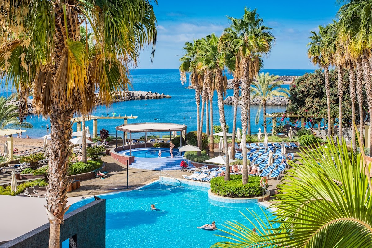 Palm trees frame the tiered pool area at Calheta Beach - All Inclusive - Savoy Signature, with bright blue water, loungers, and the Mediterranean Sea beyond.
