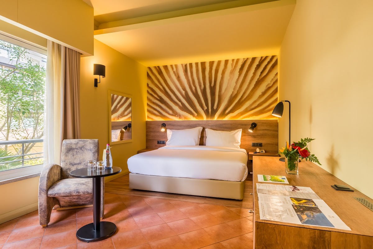 Modern resort bedroom with yellow walls, white bedding, wood headboard with striped artwork, terracotta floor, and garden views through large window.