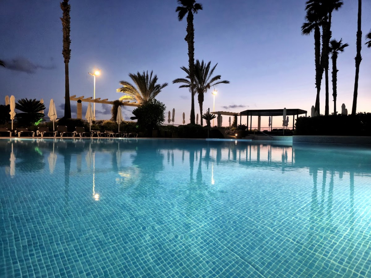Illuminated pool at dusk surrounded by tall palms, loungers, and umbrellas at Calheta Beach Savoy Signature.