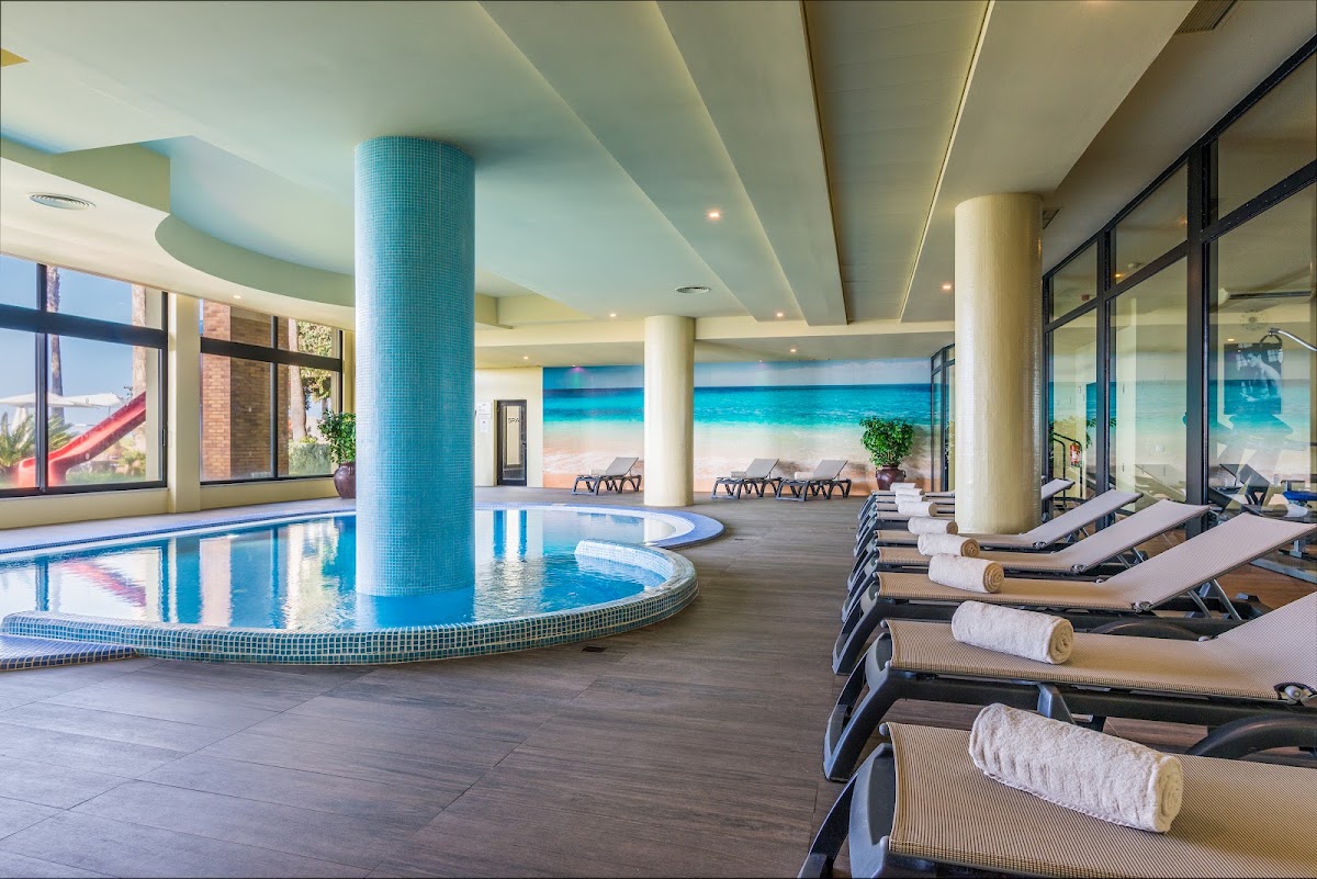 Indoor pool area with blue tile column, lounge chairs with rolled towels, and beach view through floor-to-ceiling windows at Calheta Beach resort.