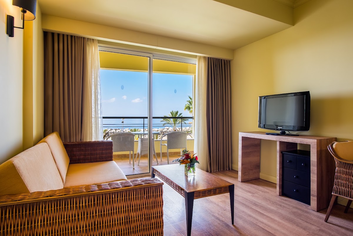 Hotel suite with yellow walls, wooden sofa, TV, and glass doors opening to a balcony overlooking the beach and ocean.