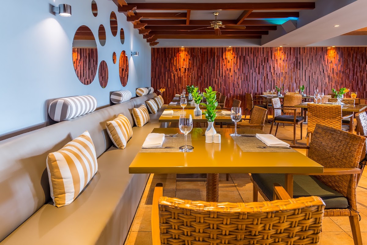 Modern dining room with yellow tables, striped cushions on banquette seating, woven chairs, and decorative wooden wall panels at Savoy Signature.