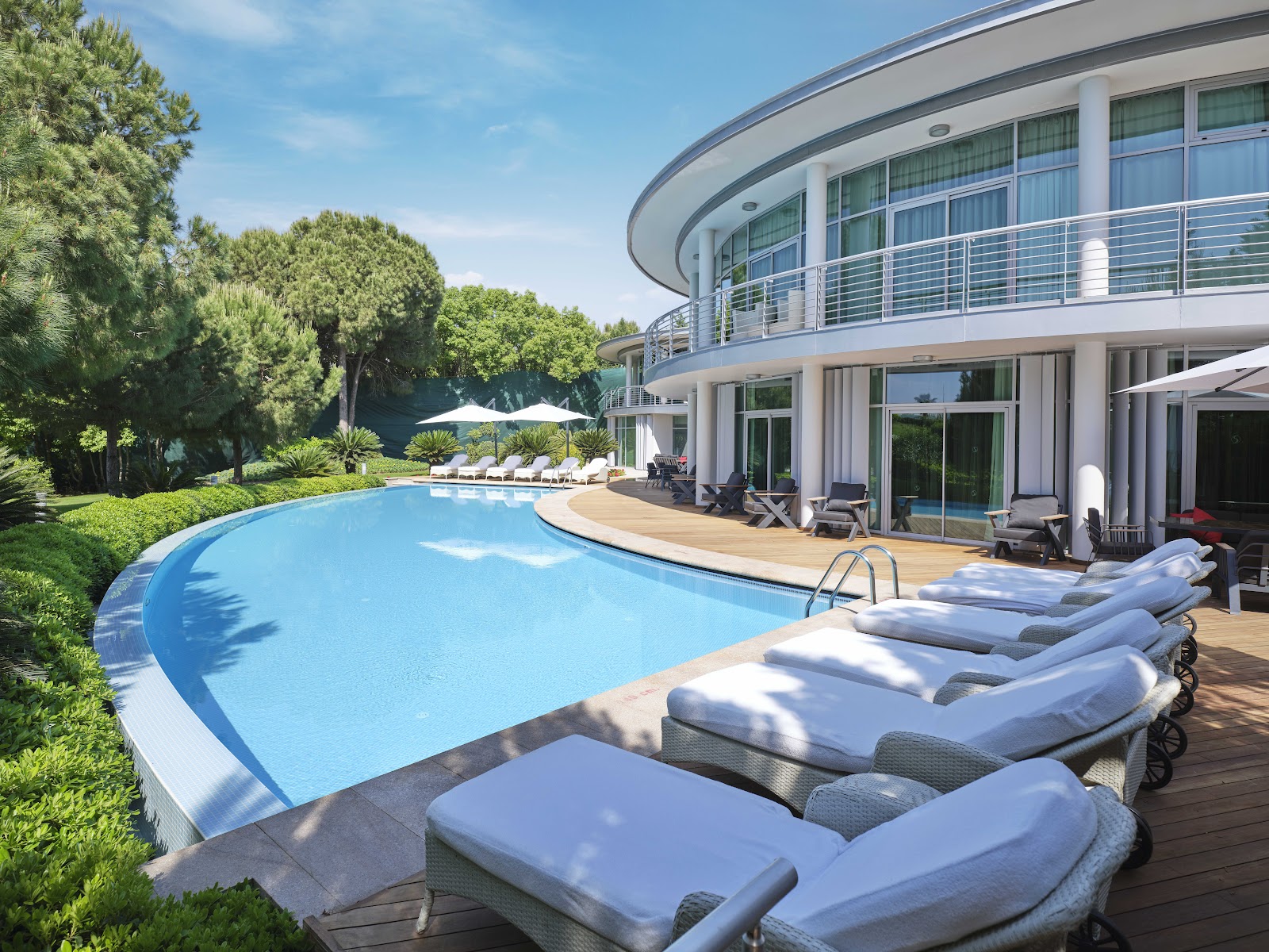 Curved white building with blue-railed balconies overlooks kidney-shaped pool with loungers on wooden deck, surrounded by pine trees under clear sky.