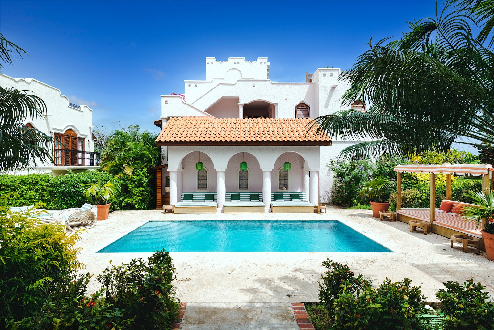 White colonial-style building with terracotta roof and arched windows overlooks a rectangular blue pool surrounded by palm trees and loungers.