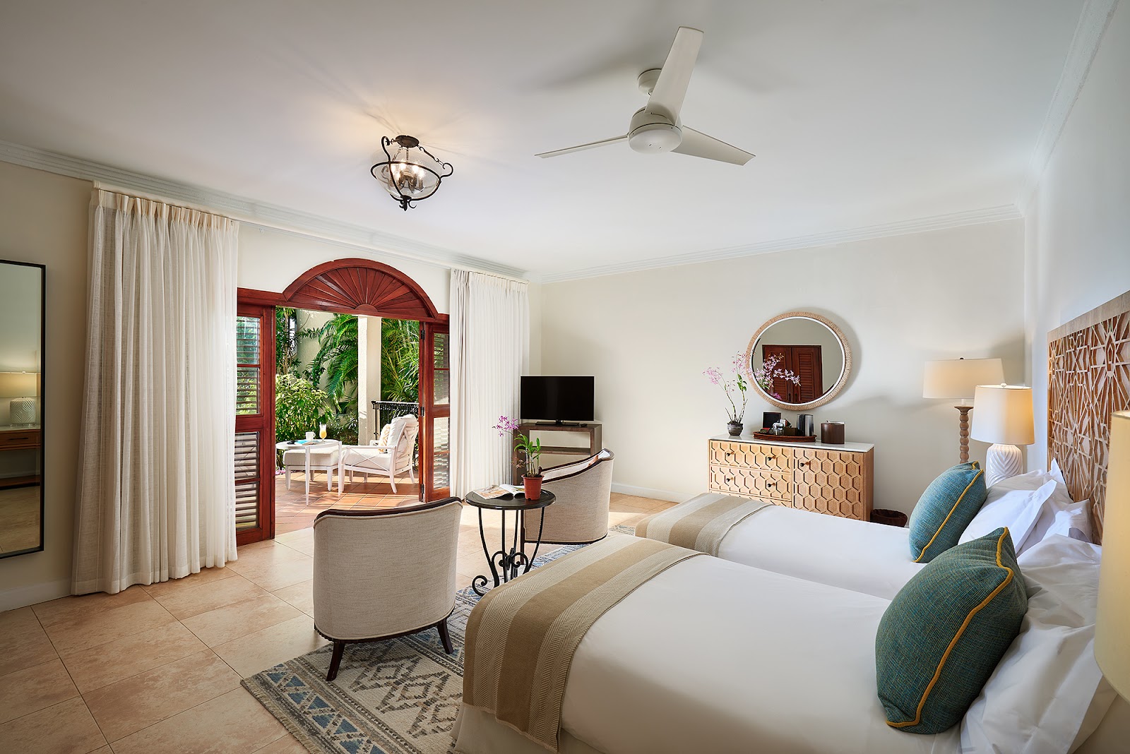 Spacious suite with two beds, sitting area, and red-framed doors opening to a tropical patio with lounge chairs at Cap Maison Resort & Spa.