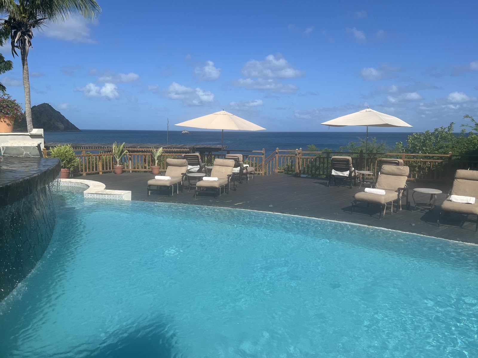 Resort pool with lounge chairs and cream umbrellas overlooking the Caribbean Sea and a green mountainous island.