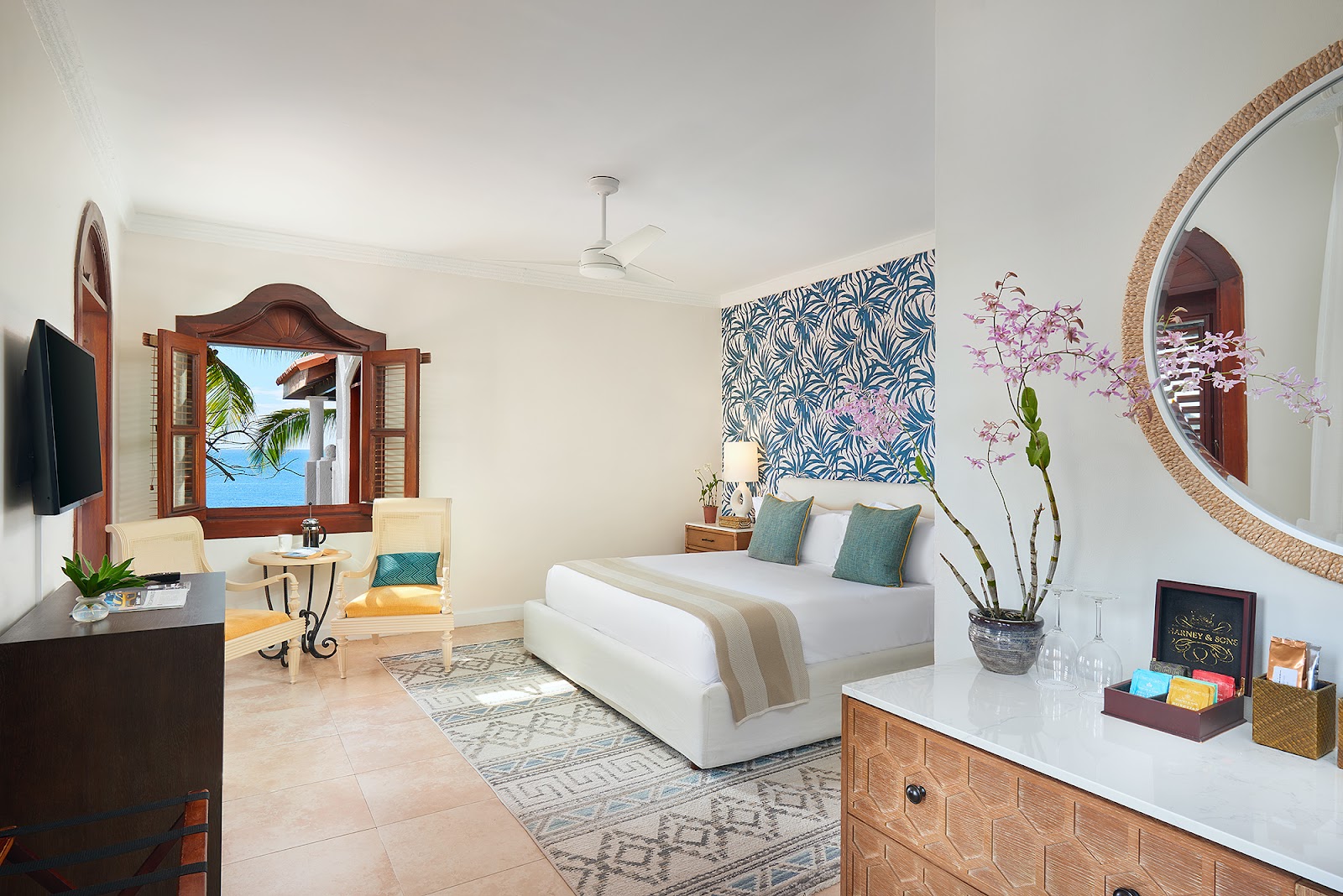 Resort suite at Cap Maison with white bed, teal pillows, ocean view through wooden-framed window, and orchid arrangement on dresser.