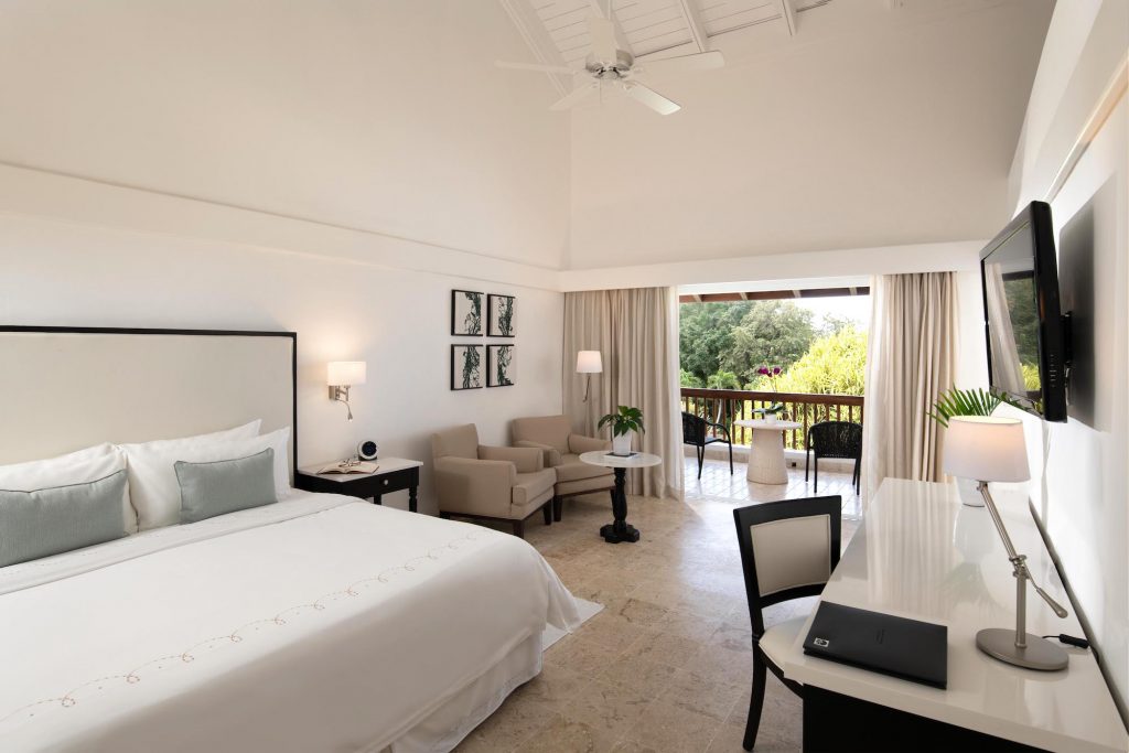 Spacious resort bedroom with king bed, sitting area, and balcony overlooking lush grounds at Casa de Campo Resort & Villas.