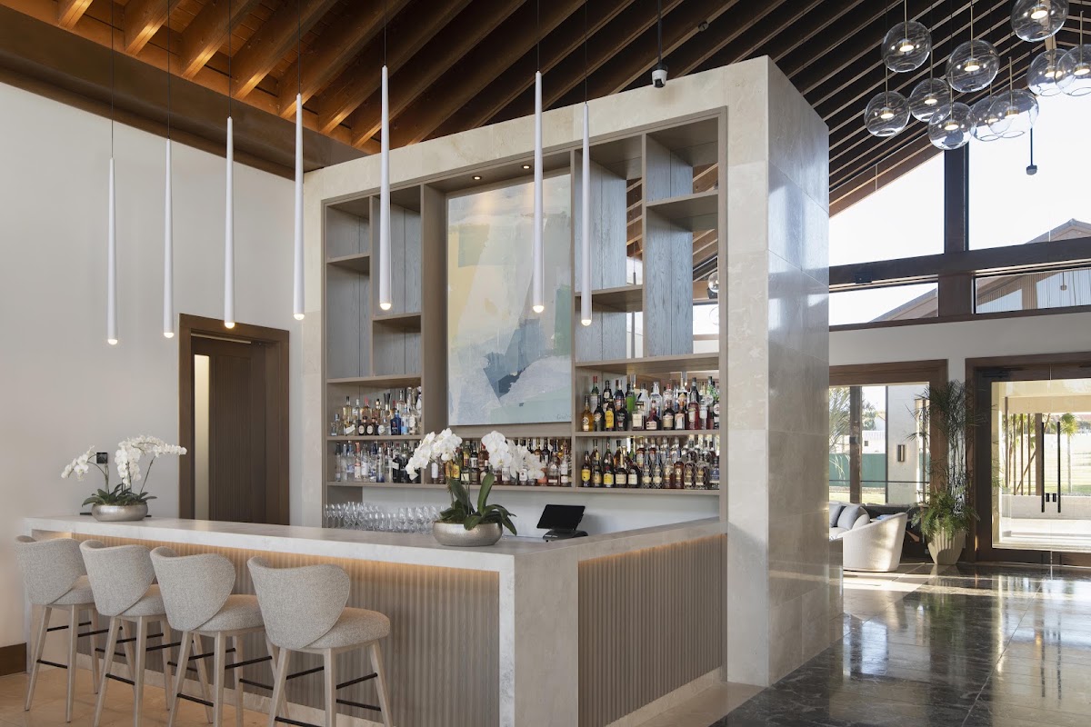 Modern bar at Casa de Campo Resort with white upholstered stools, shelves of spirits, hanging pendant lights, and exposed wood ceiling beams.