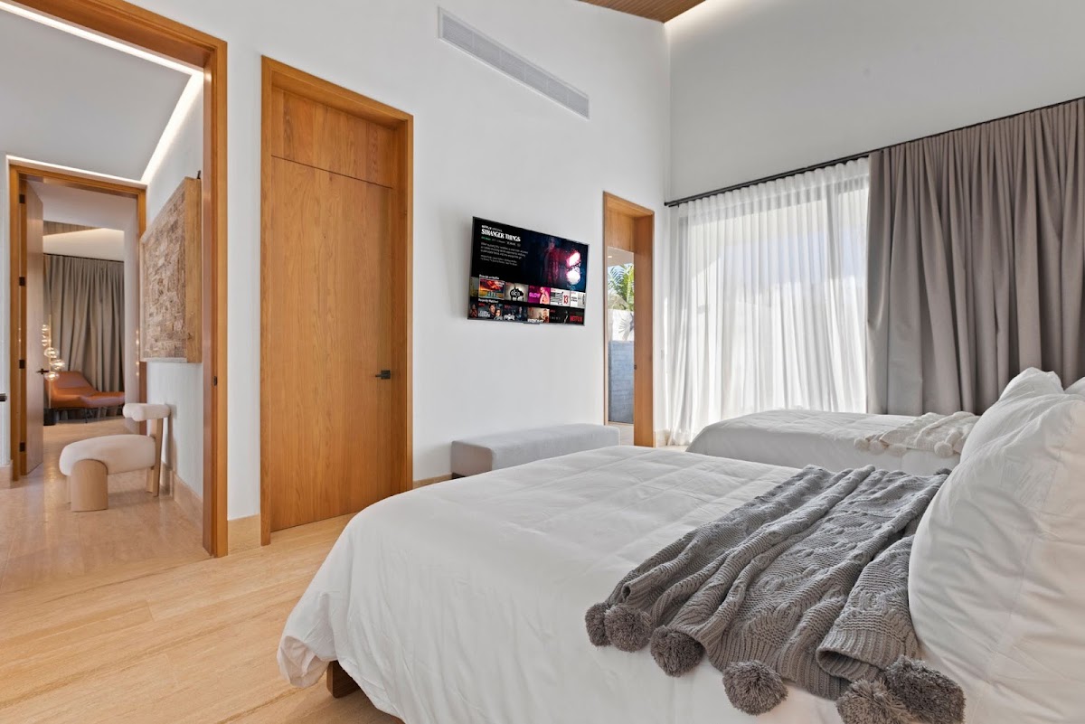 Modern resort bedroom with two beds, wooden doors, white walls, and access to a sitting area and terrace with ocean view.