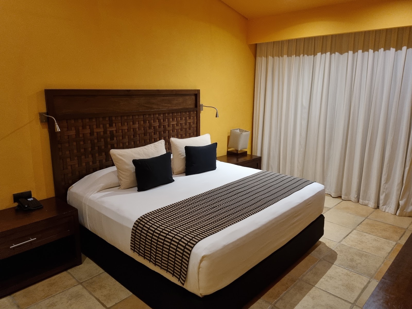 Resort bedroom with king bed, dark wood headboard, white linens, striped bed runner, and bright yellow walls with floor-to-ceiling curtains.