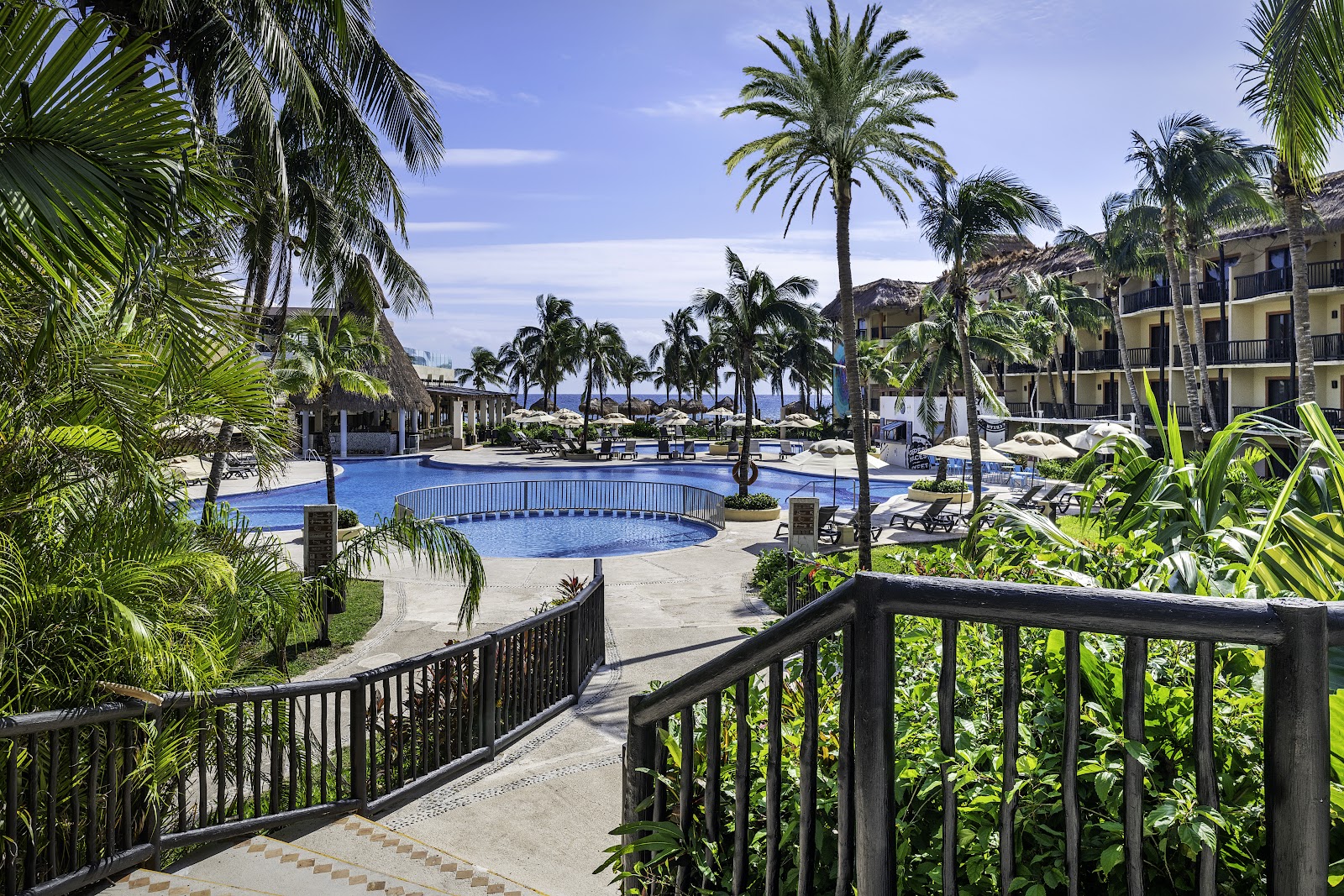 Resort pool with blue loungers, thatched-roof buildings, and palm trees under clear sky at Hotel Catalonia Riviera Maya.