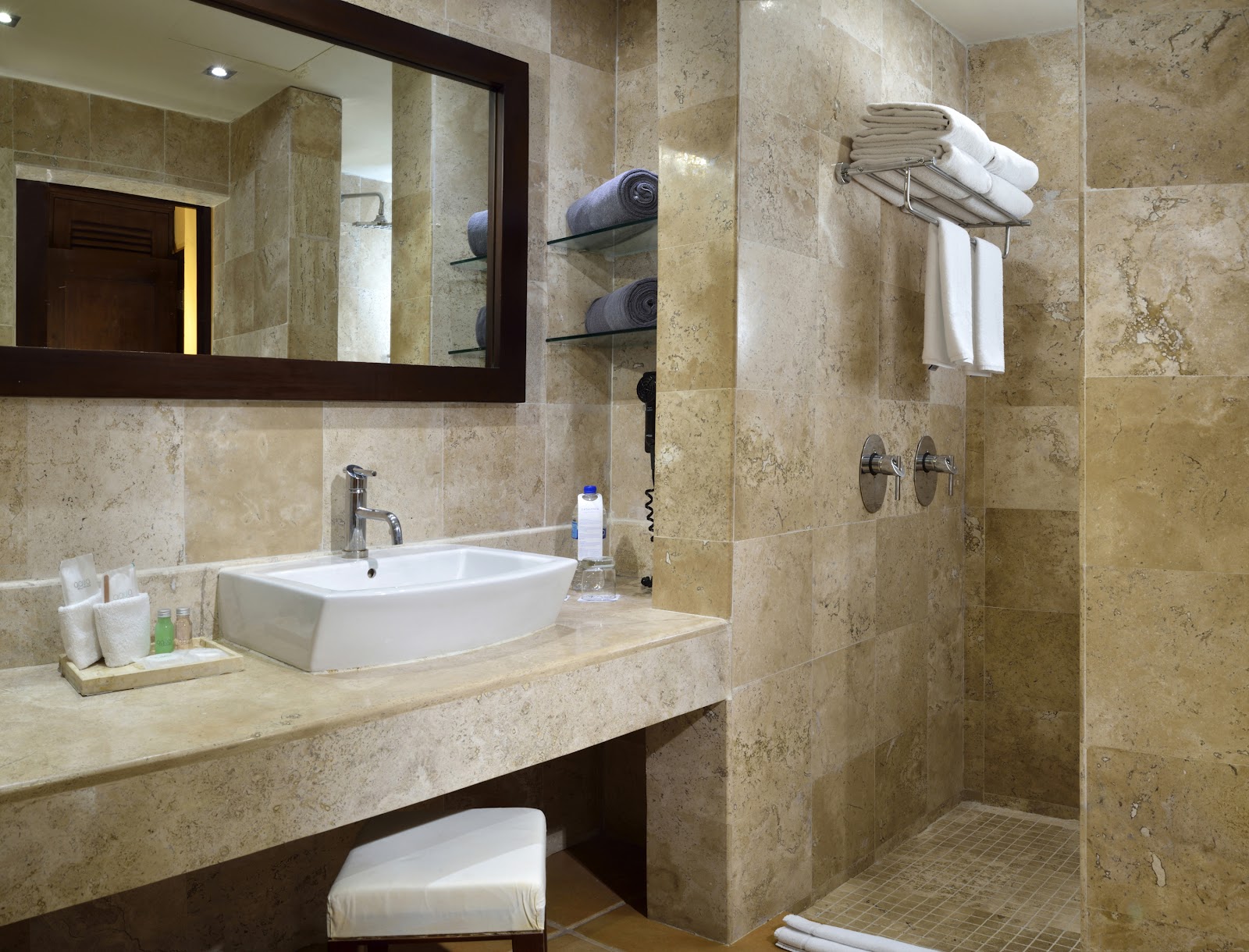 Bathroom with travertine tile walls, rectangular white sink, framed mirror, walk-in shower with towel rack, and rolled towels on shelves.