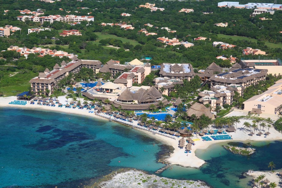Aerial view of an all-inclusive resort on a white sand beach with turquoise water, multiple tan buildings with thatched roofs, blue pool areas, and lush green hills beyond.