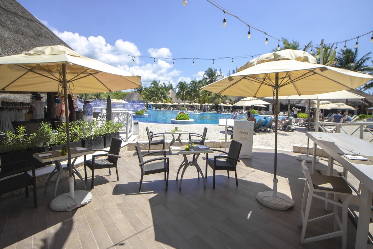 Shaded dining area with cream umbrellas overlooking a large resort pool surrounded by loungers and palm trees under blue sky.