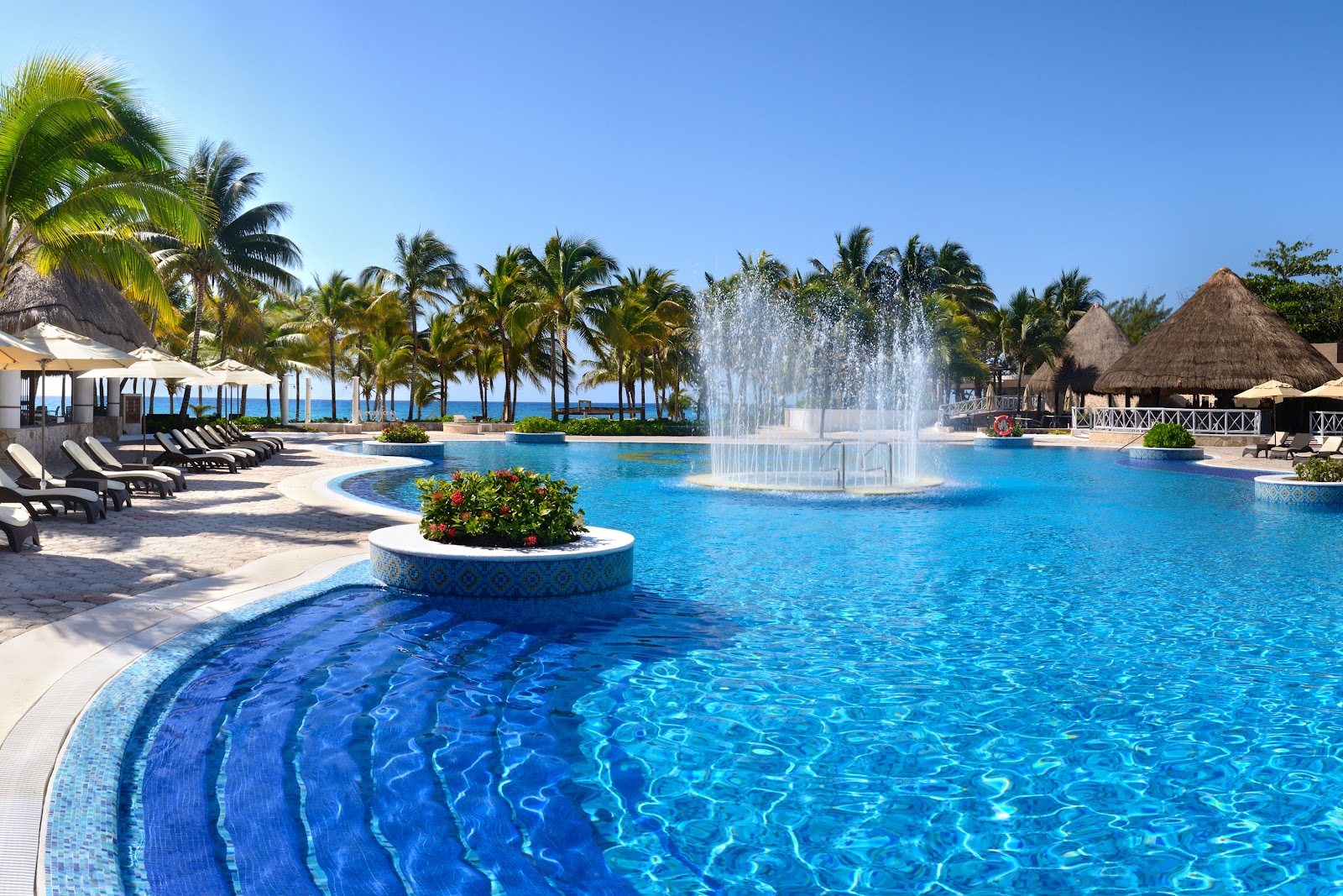 Resort pool with thatched-roof bar and loungers overlooking turquoise Caribbean Sea and white sand beach lined with palm trees.