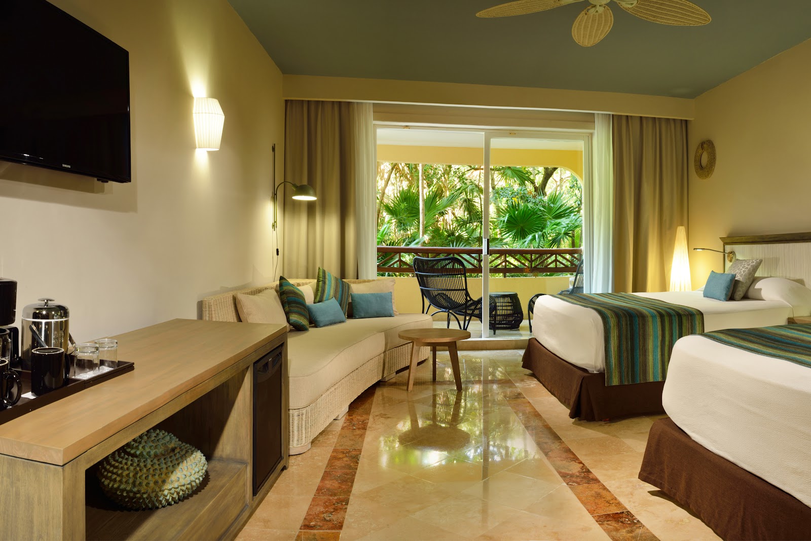 Resort room with two beds, seating area, and balcony overlooking tropical palms and vegetation.