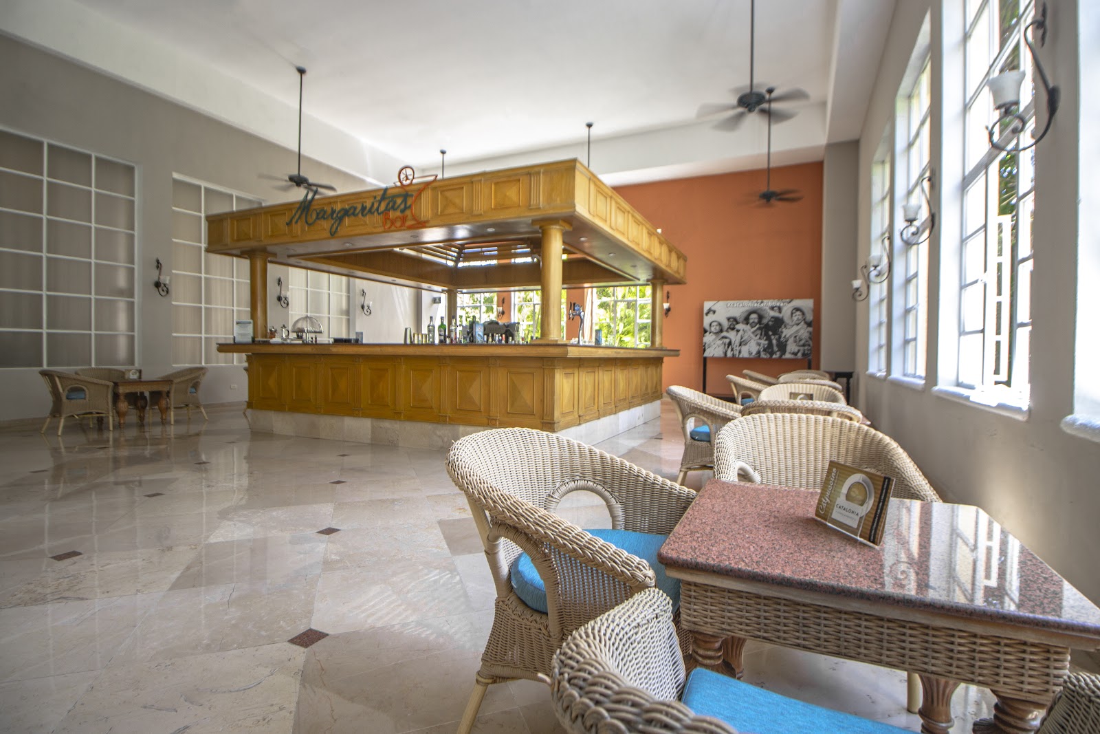 Wicker chairs at tables in the airy lobby bar of Hotel Catalonia Royal Tulum, with a wooden counter and orange accent wall.