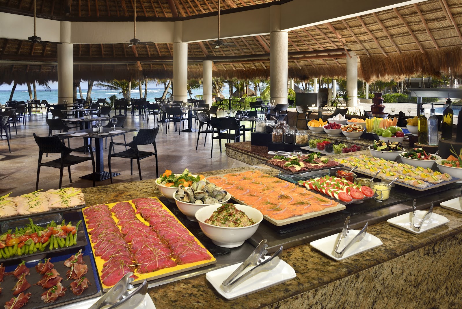 Spacious lobby with thatched roof, wooden support beams, modern seating areas with orange and blue accents, and hanging metal sculptures.