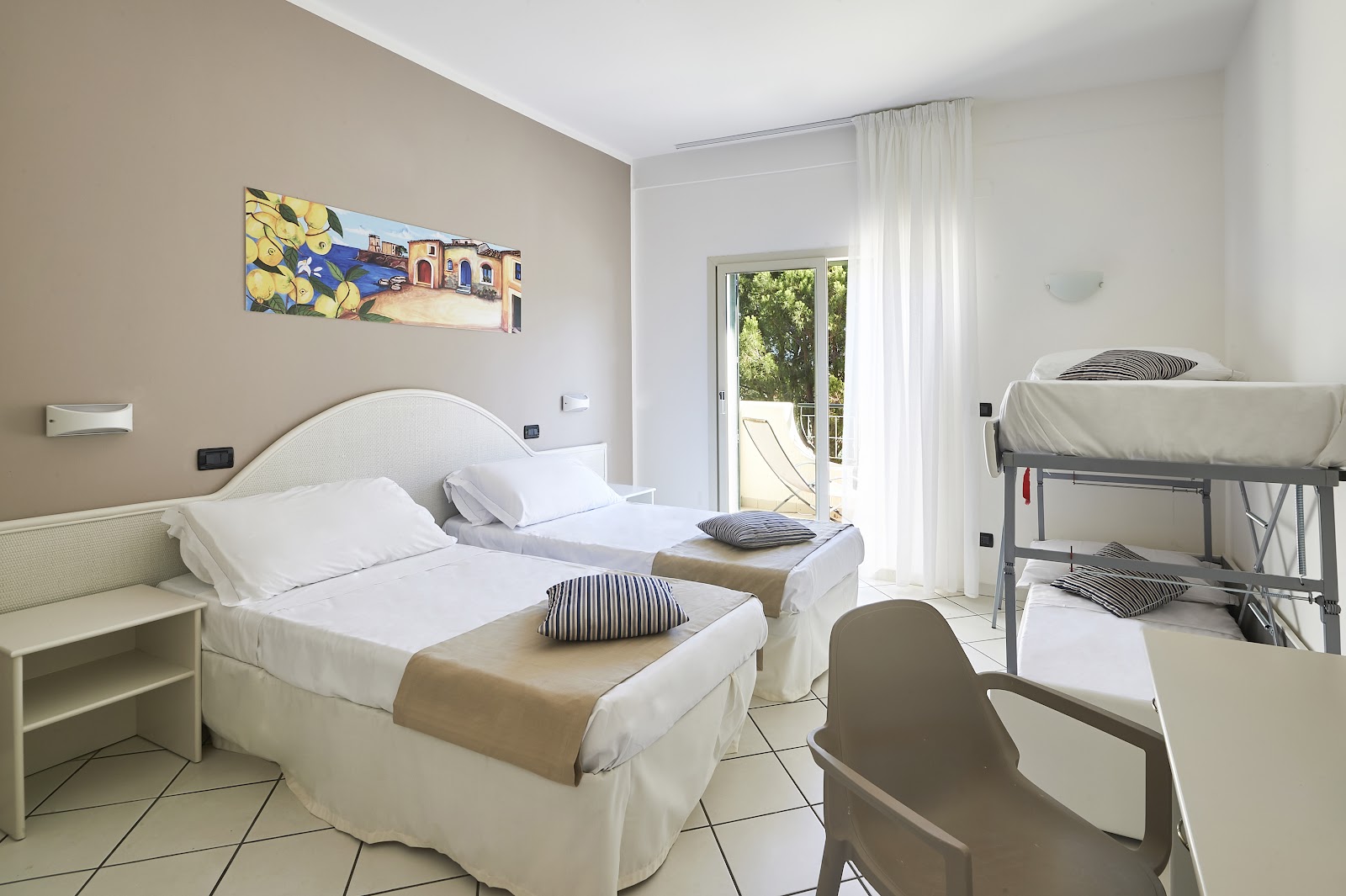 Resort room with two twin beds, bunk beds, cream and white furnishings, and a glass door opening to a balcony with greenery visible.