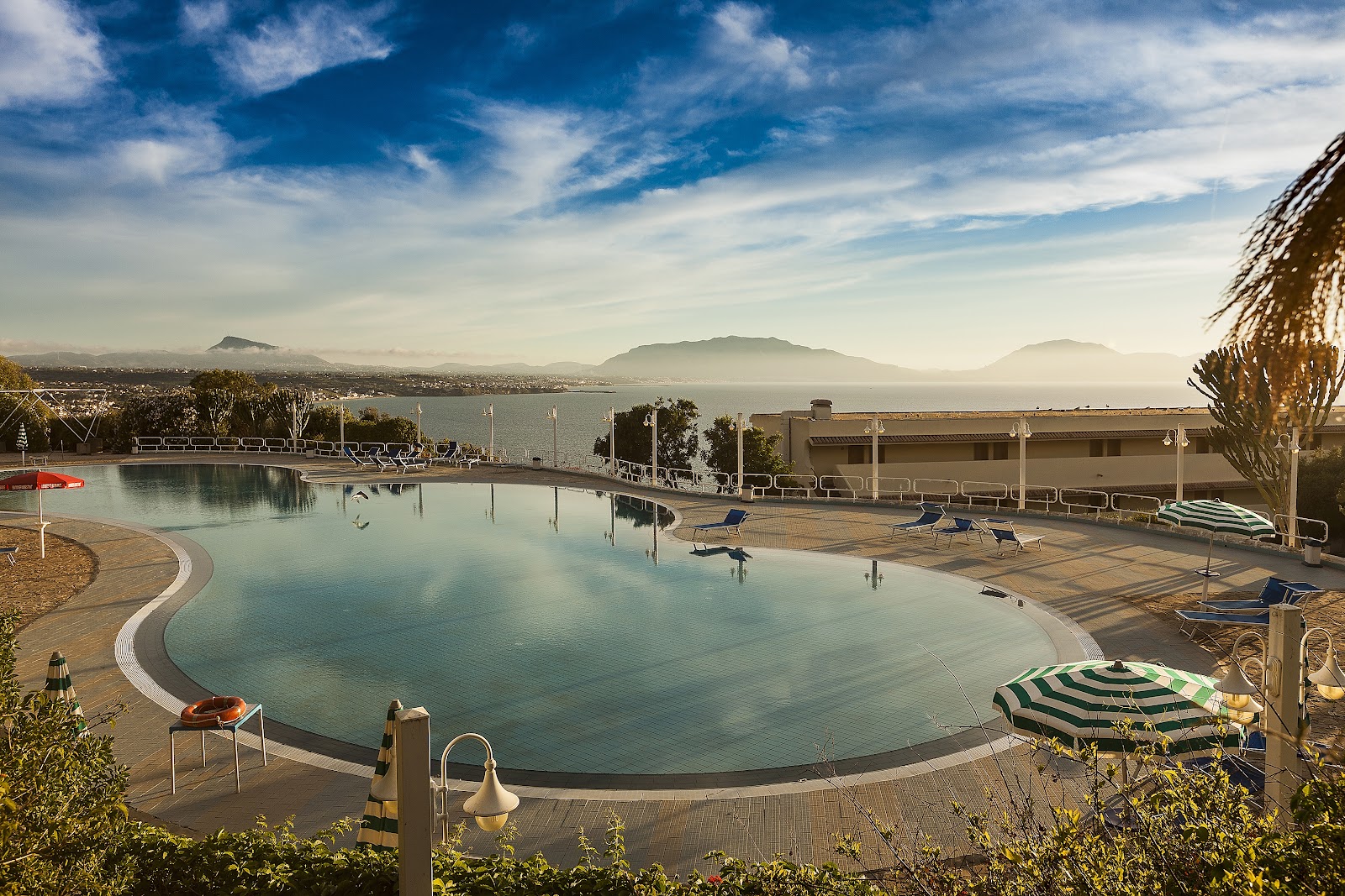 Resort pool with turquoise water, lounge chairs, and striped umbrellas overlooking a bay with mountains in the distance under blue sky.