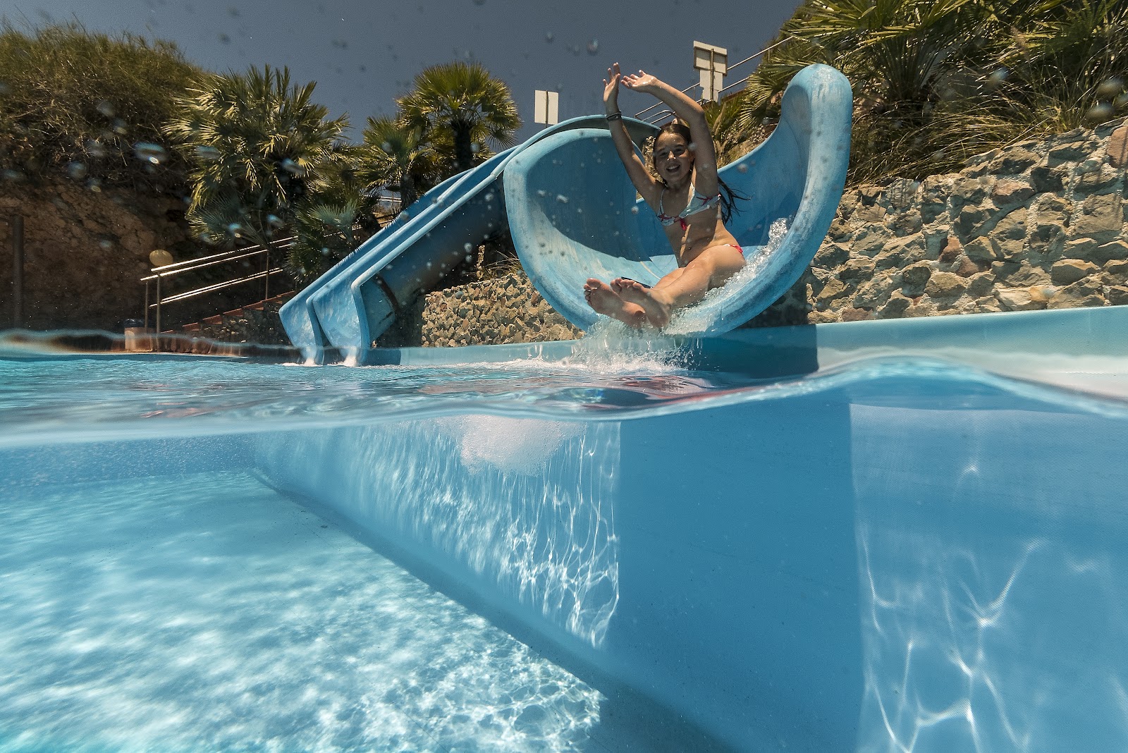 A woman slides down a blue water slide into a pool at CDSHotels Terrasini, surrounded by palm trees and sandy terrain.