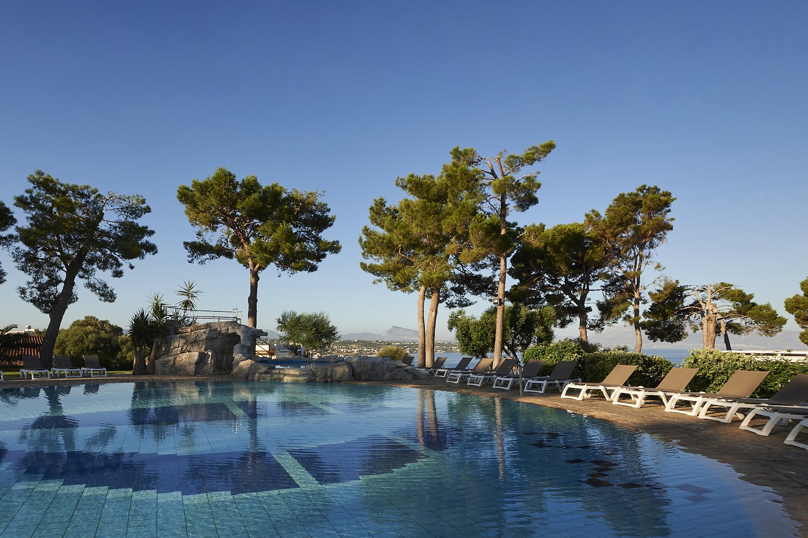 Resort pool with white loungers, tall pine trees, and a rocky waterslide feature overlooking the Mediterranean Sea.