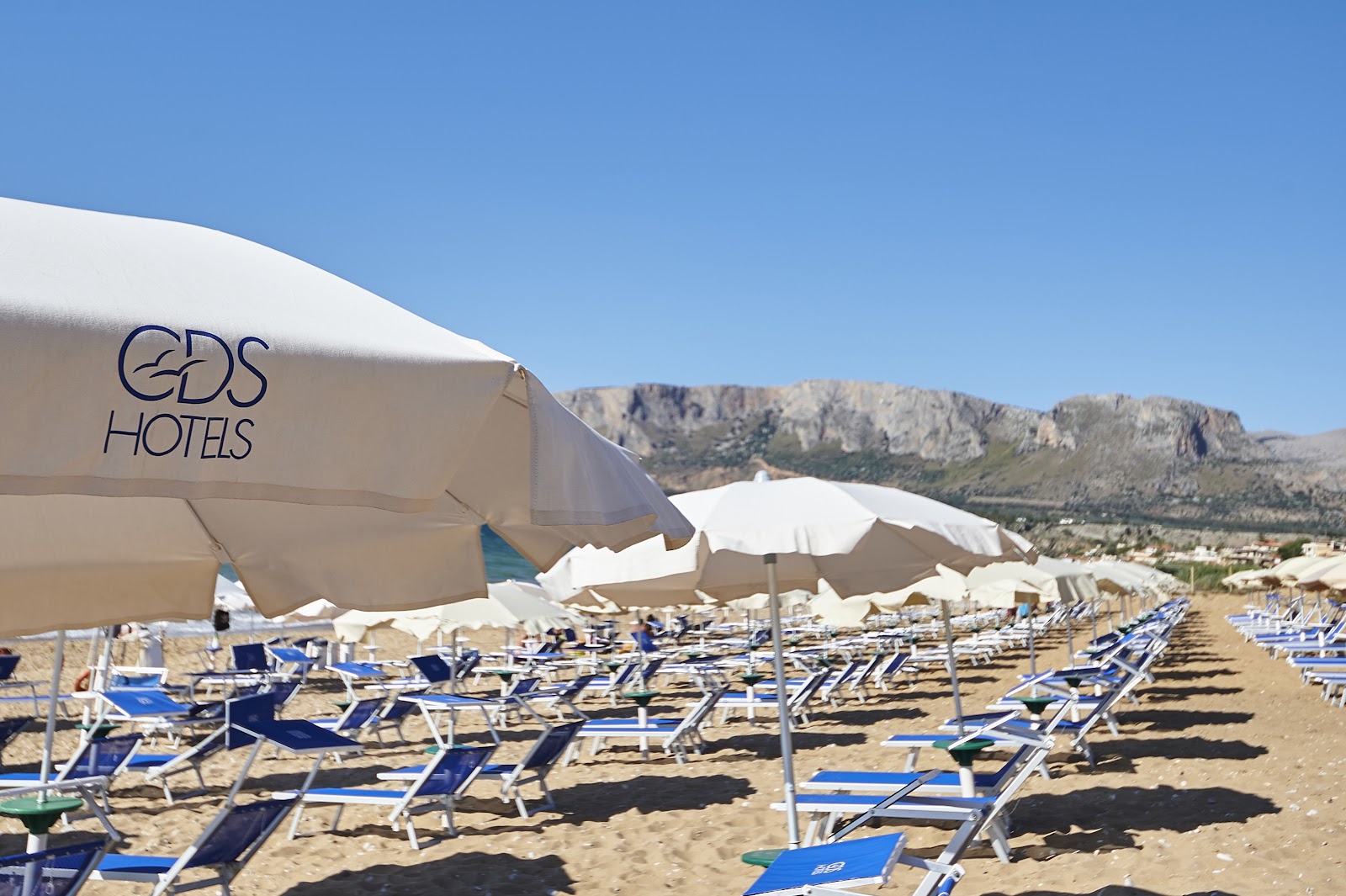 EDS Hotels beach with rows of blue loungers, white umbrellas, and rocky mountains in the background under clear sky.