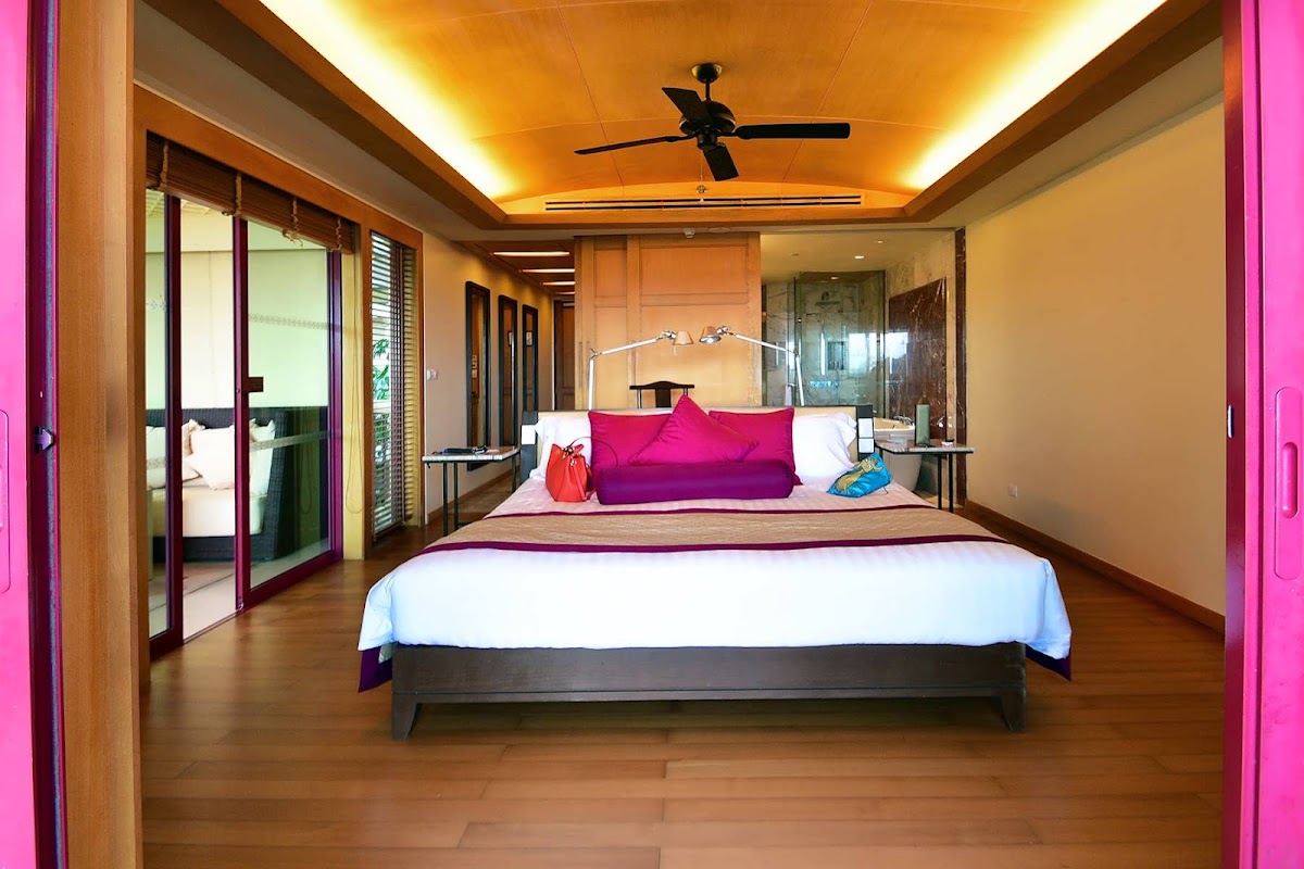Spacious resort bedroom with wooden floors, large bed with white linens, bright pink accents, ceiling fan, and ensuite bathroom visible through glass doors.