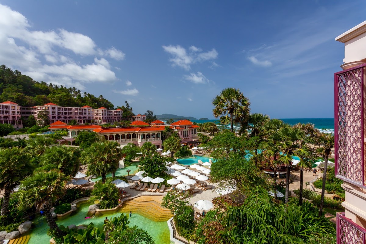 Centara Grand Beach Resort Phuket overlooks multiple pools with white umbrellas, palm trees, and the turquoise ocean beyond forested hillsides under blue sky.