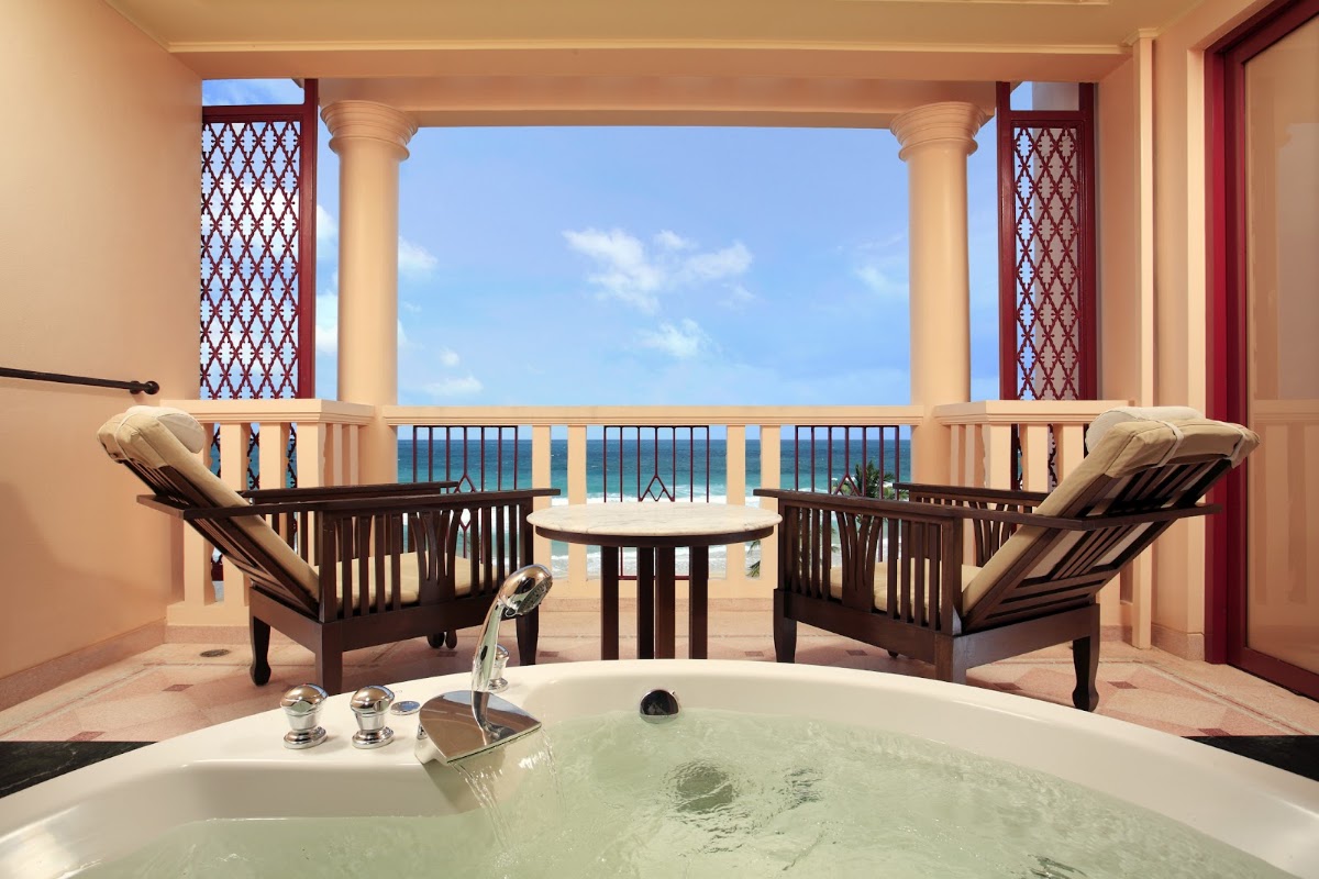 Soaking tub in foreground with two lounge chairs and small table on a beachfront balcony overlooking turquoise water under clear sky.