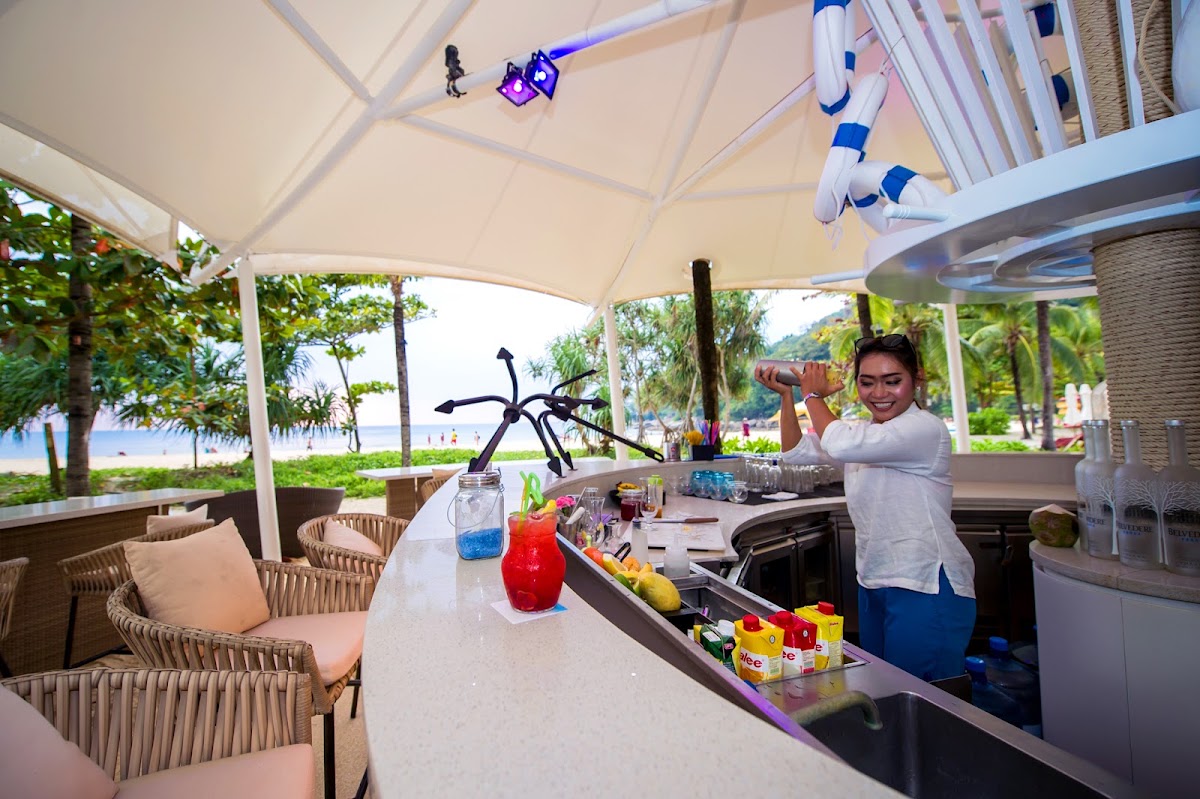 Bartender mixing a red cocktail at an outdoor beachfront bar under a white canopy, with palm trees and ocean visible beyond.