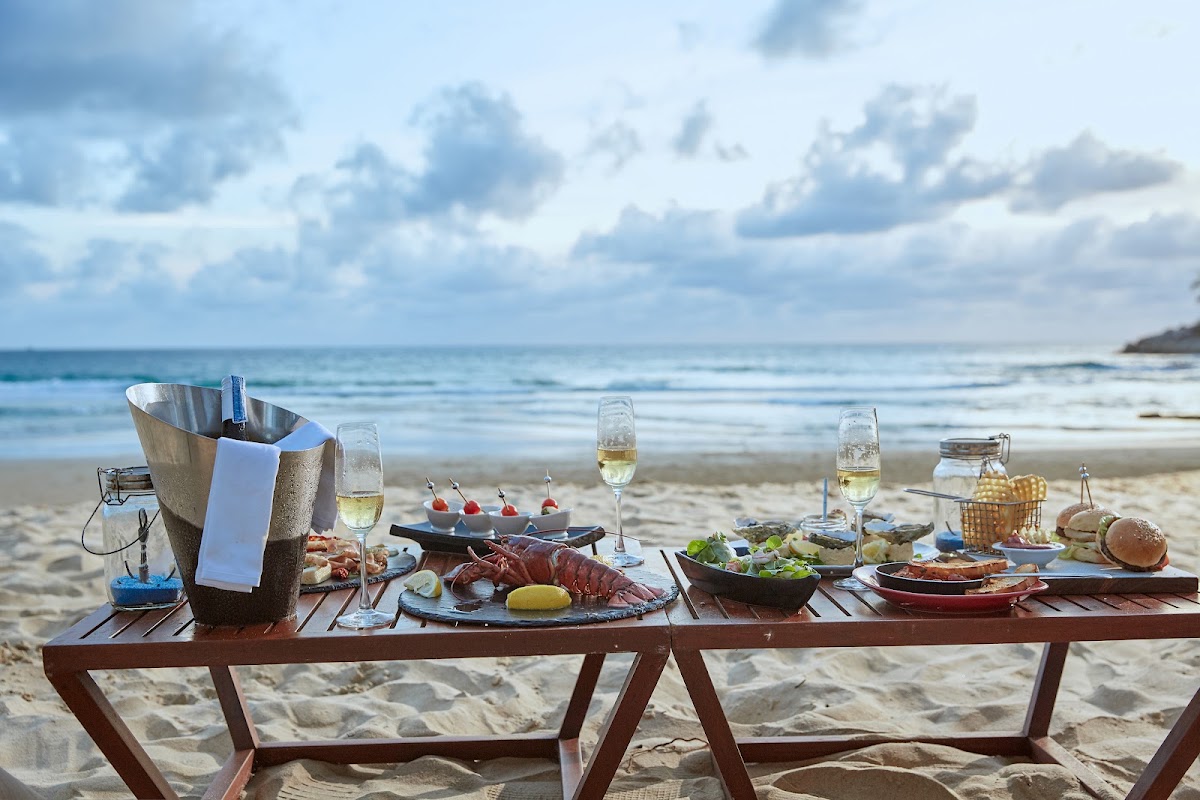 Beachfront dining table set with seafood, wine glasses, and an ice bucket on the sand at Centara Grand Beach Resort Phuket.