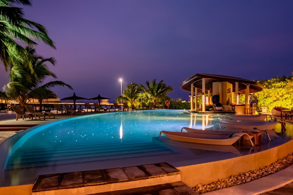 Illuminated pool at dusk with loungers, a pavilion, palm trees, and ocean views at Centara Ras Fushi Resort & Spa Maldives.