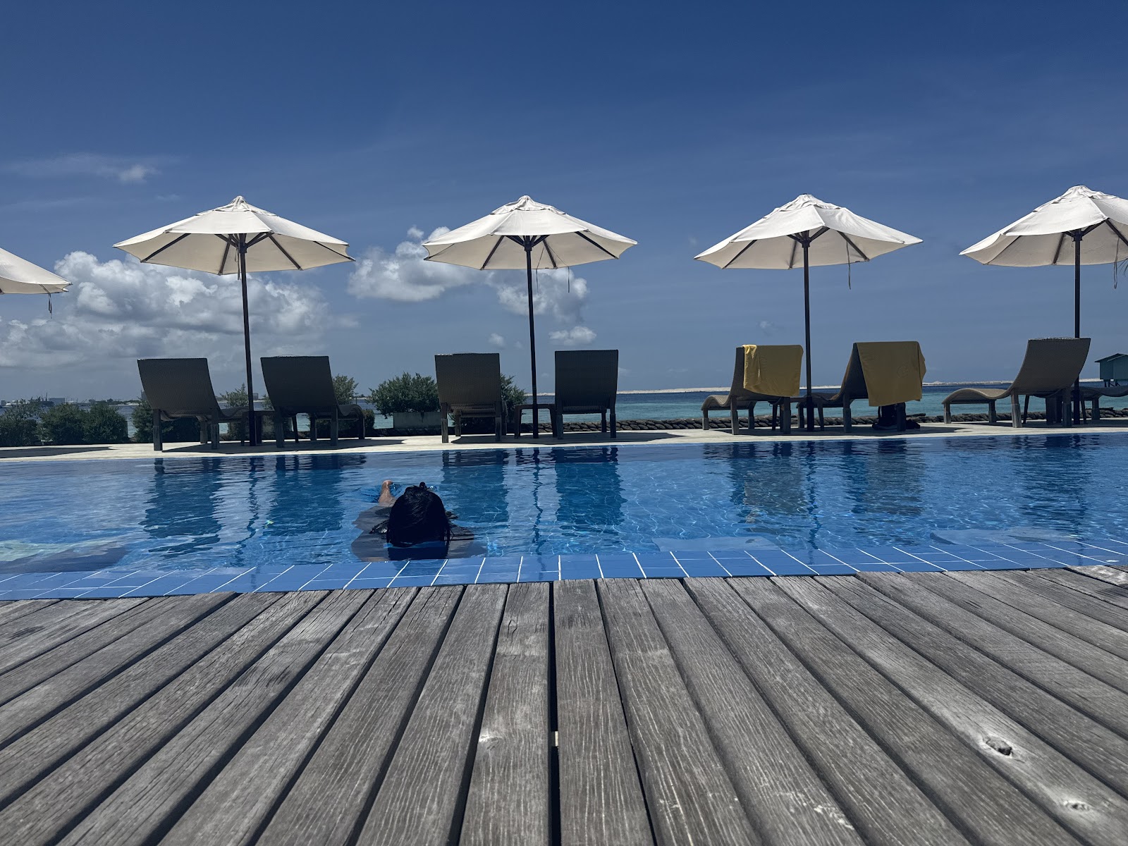 A guest floats in a resort pool with wooden deck, white umbrellas, and lounge chairs overlooking turquoise ocean water at Centara Ras Fushi.
