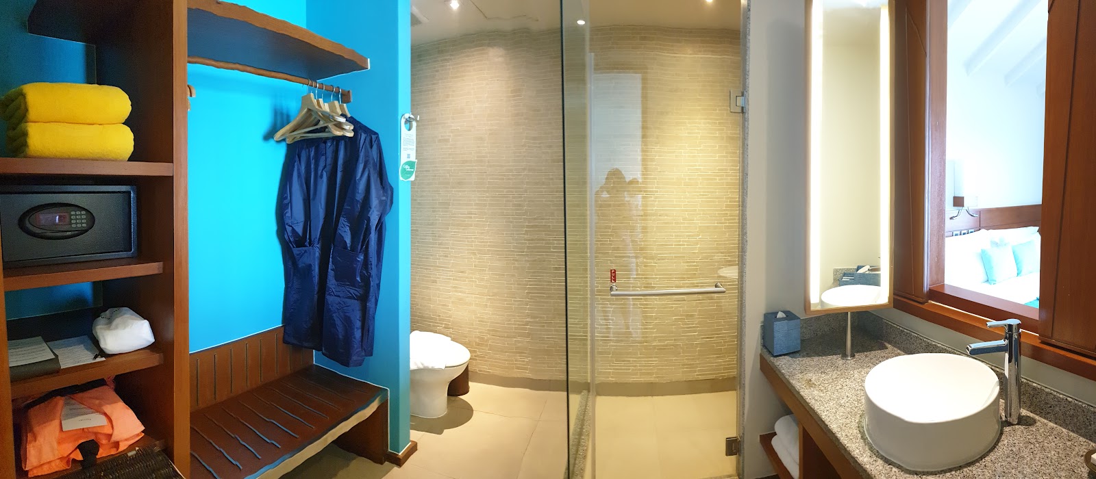 Centara Ras Fushi Resort bathroom with glass shower enclosure, round white sink, wooden shelving holding yellow towels and colorful robes, and bright blue accent wall.