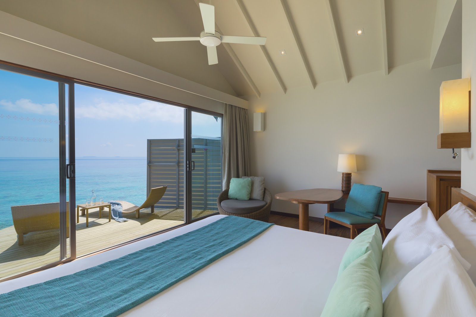 Overwater bungalow bedroom at Centara Ras Fushi with bed, seating area, and ocean view through glass doors opening to a deck.