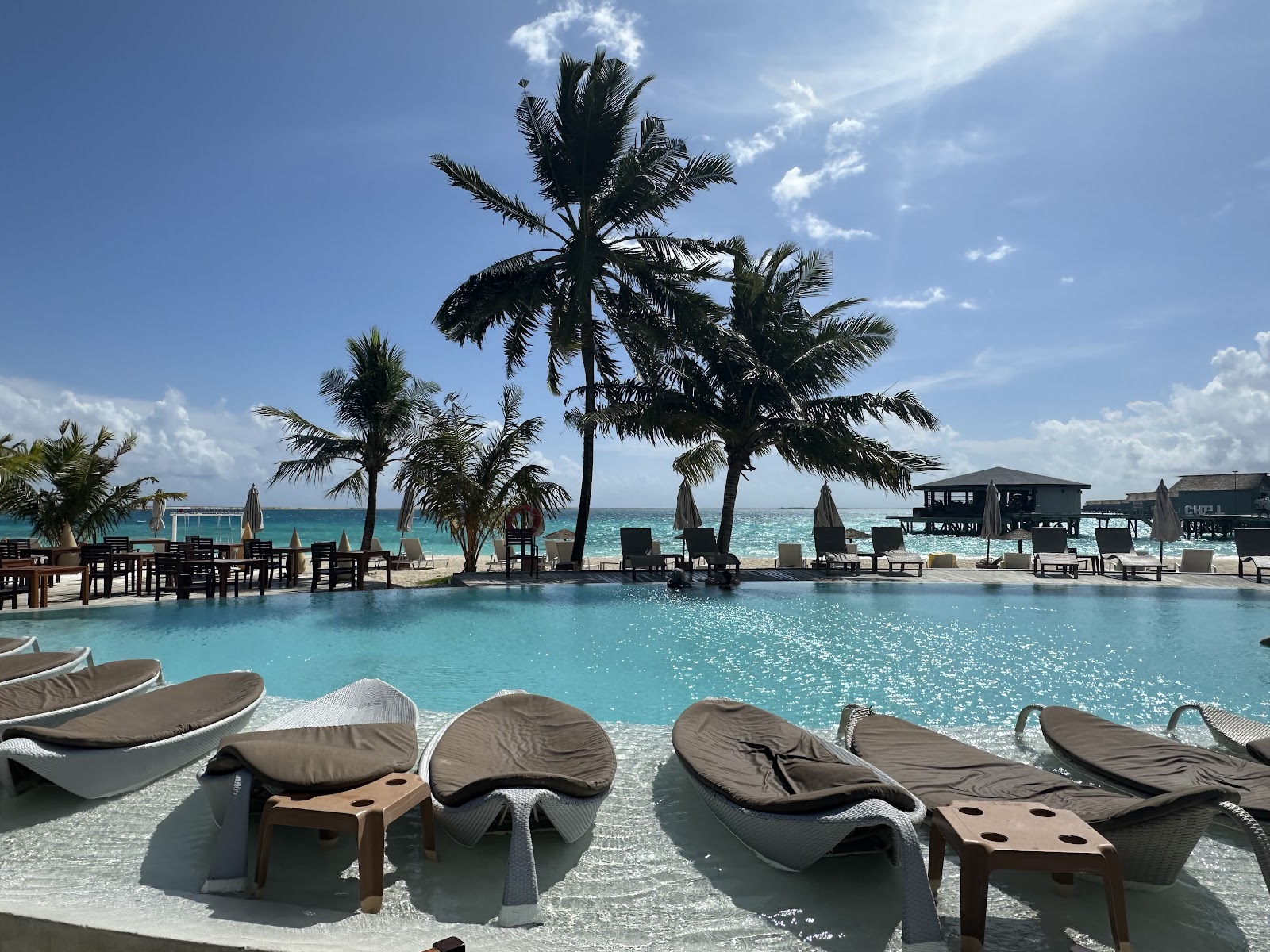 Resort pool with loungers overlooking turquoise ocean, palm trees, dining area, and beach under blue sky with white clouds.