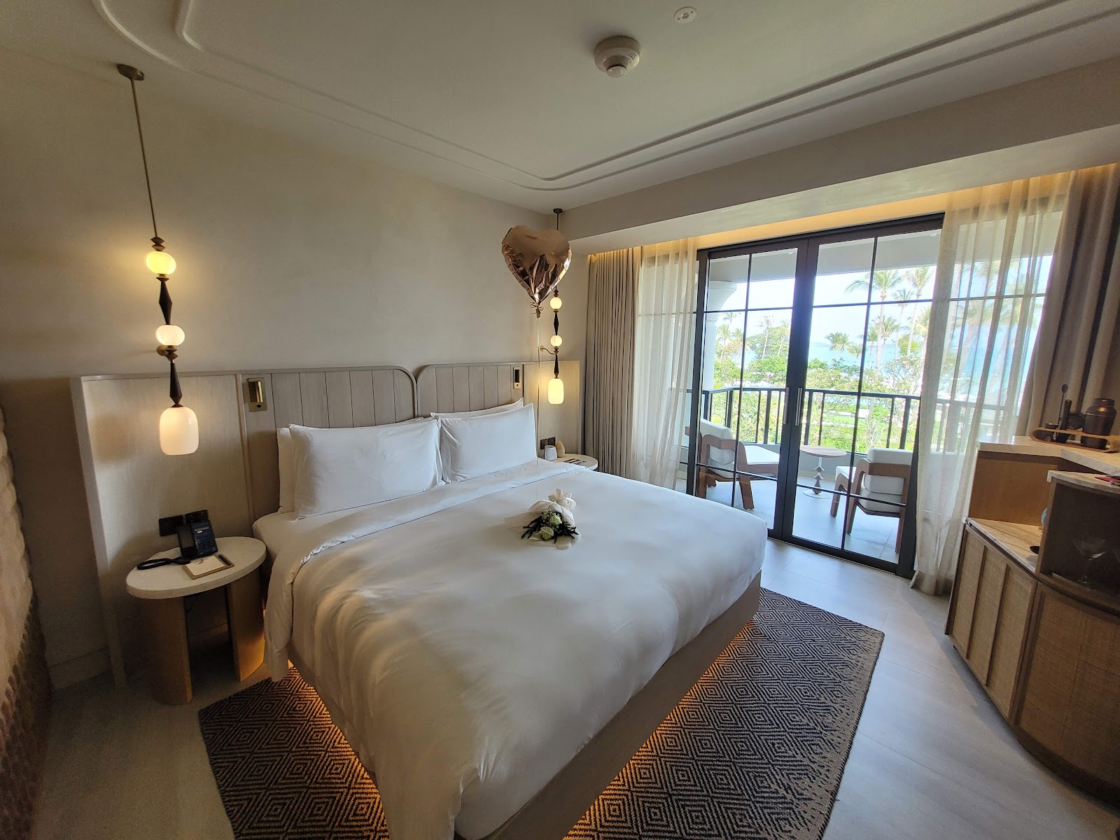 Modern resort bedroom with king bed, pendant lights, wooden furnishings, and ocean view from balcony with palm trees visible.