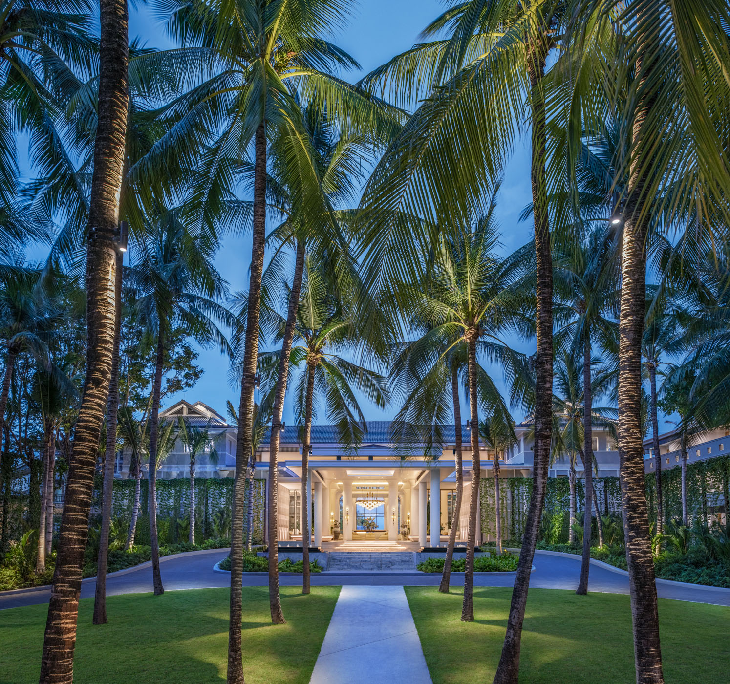 Palm-lined walkway leading to an illuminated pavilion entrance at dusk, with manicured lawns flanking the path at Centara Reserve Samui.
