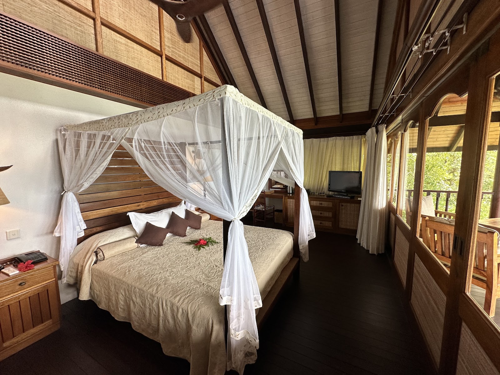 Wooden bungalow bedroom with four-poster bed draped in white mosquito netting, TV, and glass doors opening to a deck.