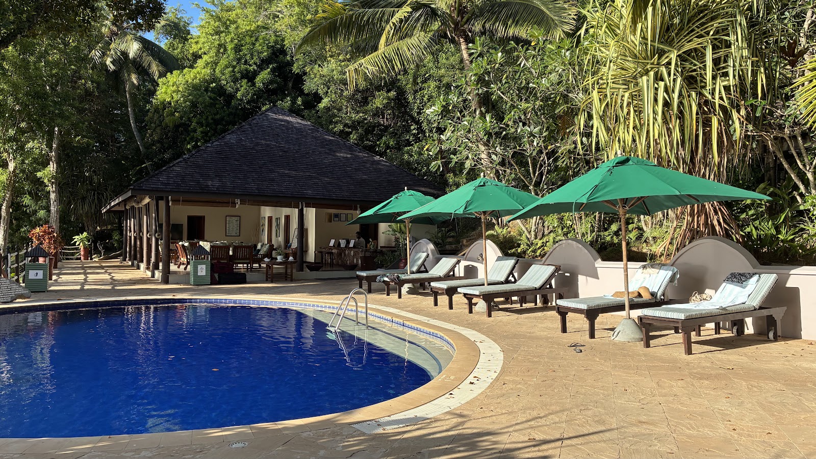 Circular pool with blue water bordered by loungers under green umbrellas, pavilion with dark roof, and dense tropical vegetation at Cerf Island Resort.