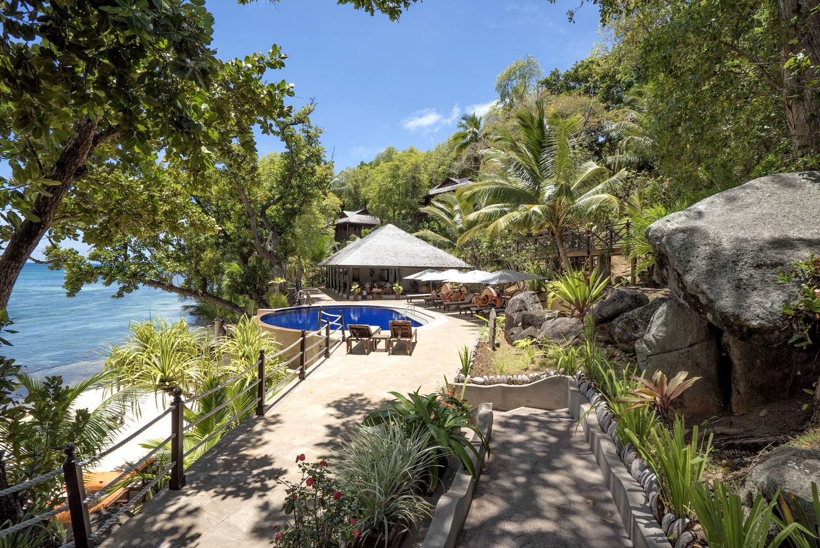Wood-beamed spa treatment room with massage bed in foreground, two lounge chairs, and floor-to-ceiling windows overlooking tropical forest at Cerf Island Resort.