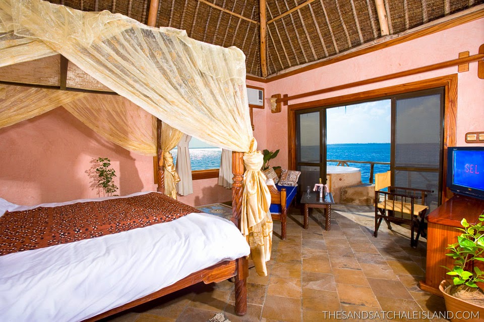 Resort bedroom with four-poster canopy bed, pink walls, thatched roof, and ocean view through glass doors.