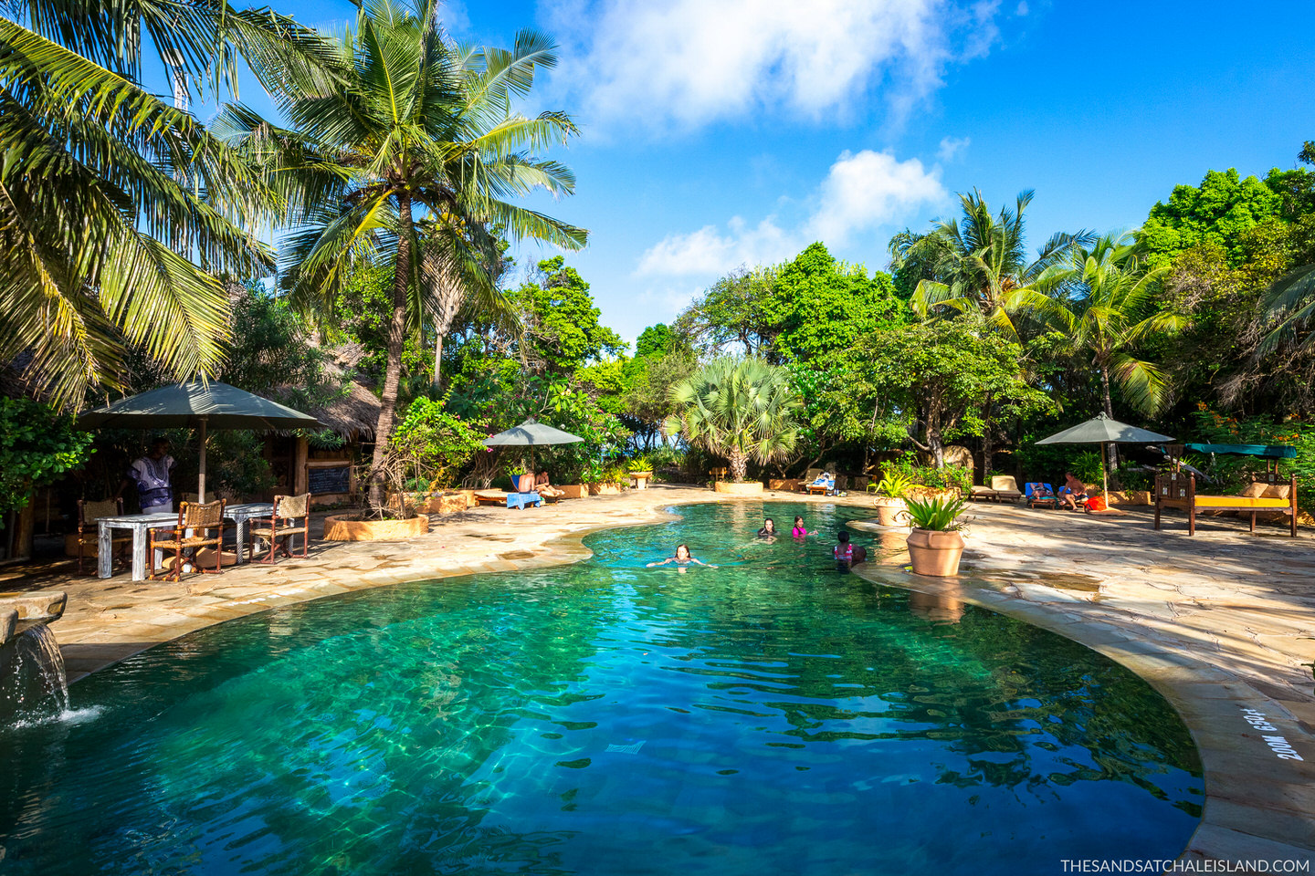 Clear lagoon pool surrounded by palm trees and tropical vegetation with swimmers, umbrellas, and thatched structures at Chale Island Resort.