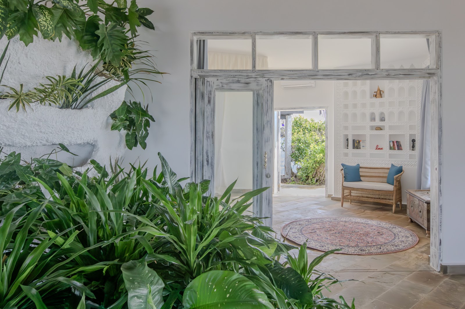 Indoor plants frame the entrance to a bright suite with whitewashed walls, rattan seating with blue cushions, and open doors leading to a garden courtyard.