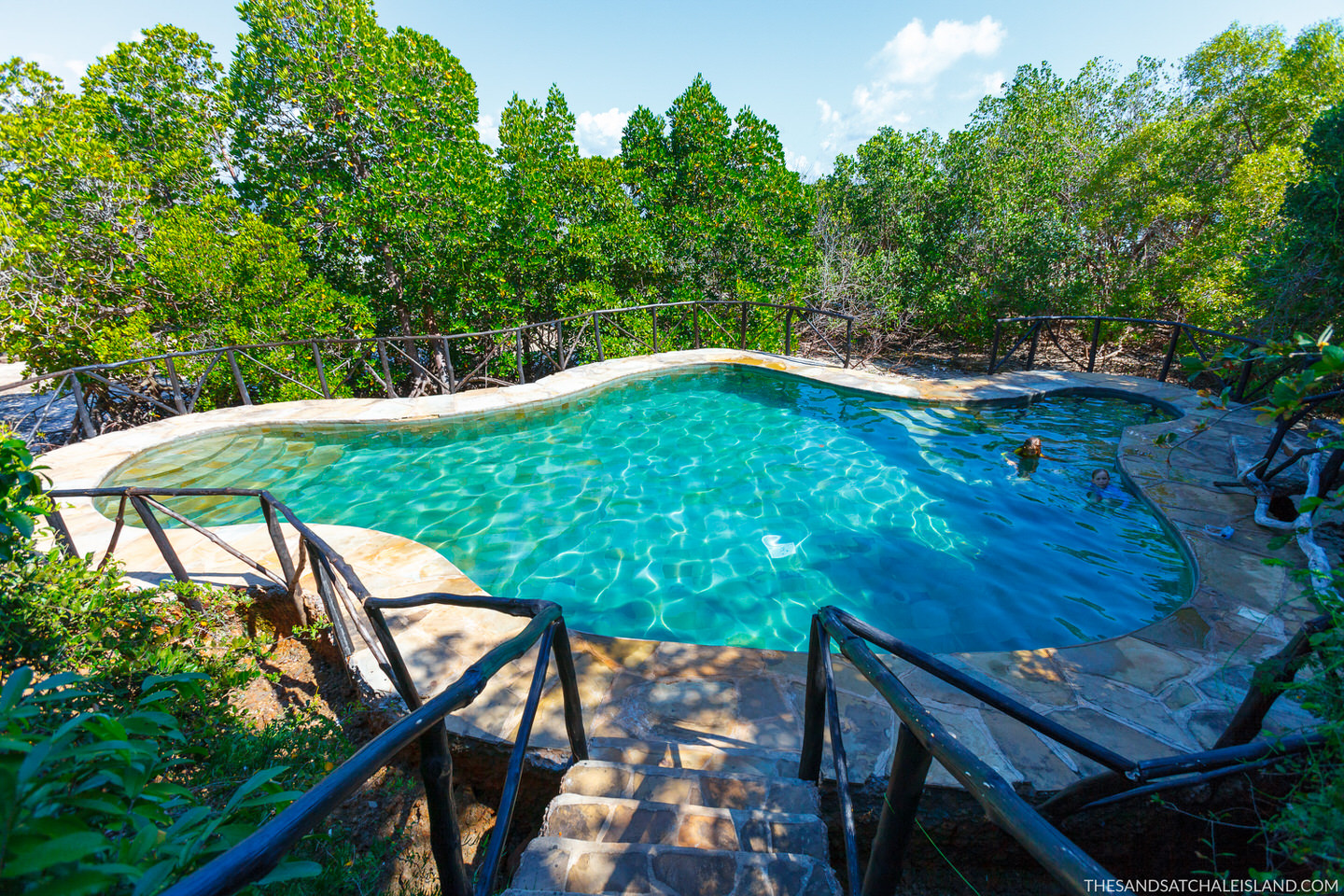 Aerial view of thatched-roof bungalows in turquoise lagoon waters beside dense green island vegetation under clear blue sky.