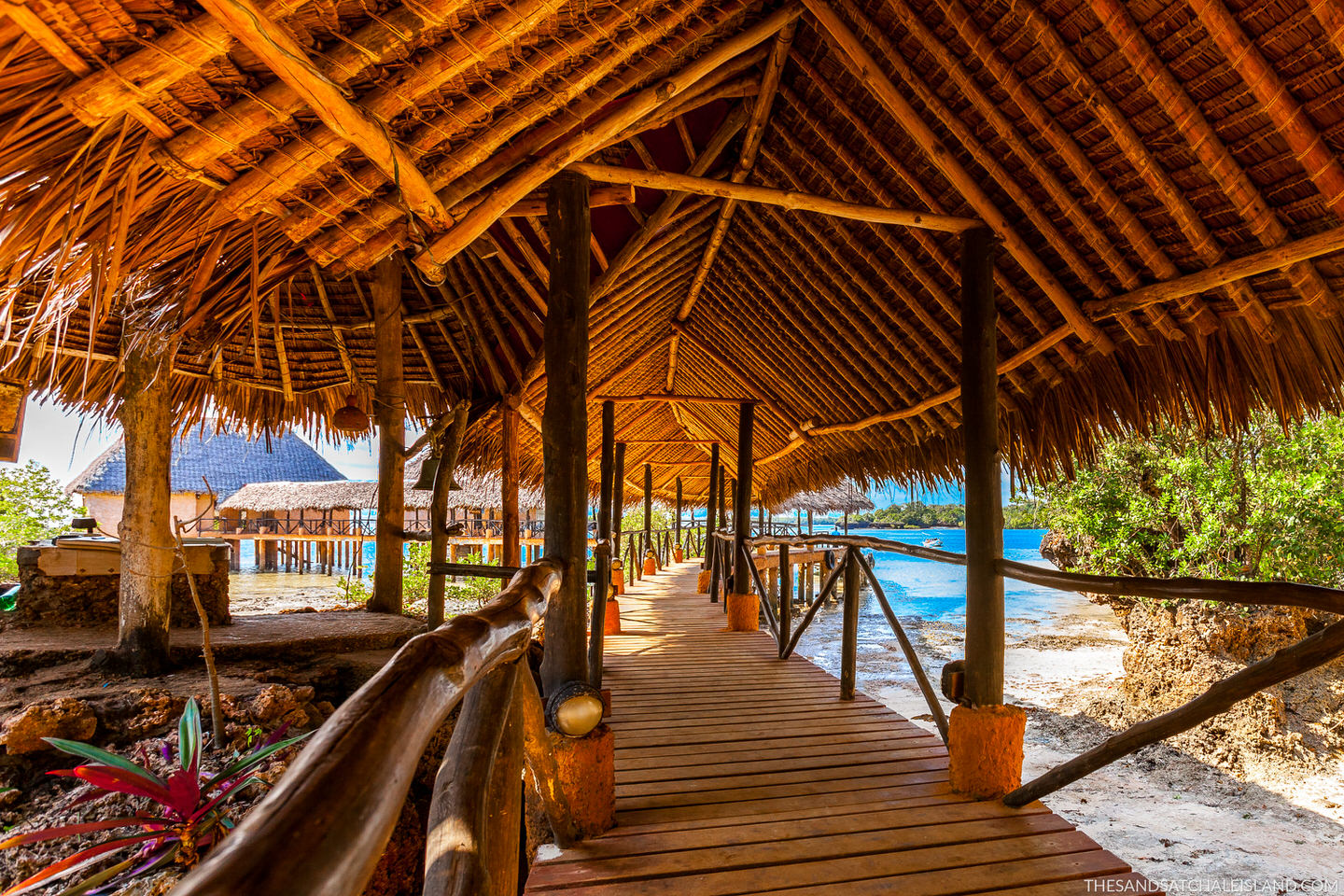 Wooden boardwalk with thatch-roof structures leading toward turquoise ocean water at a beachfront resort.
