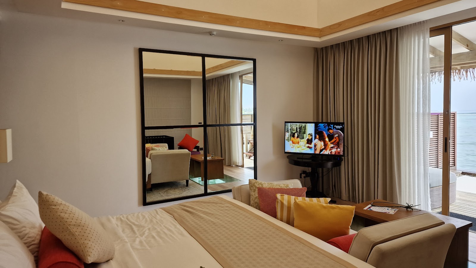 Resort suite with bed, seating area, TV, and ocean view through glass doors to a deck at Cinnamon Hakuraa Huraa Maldives.